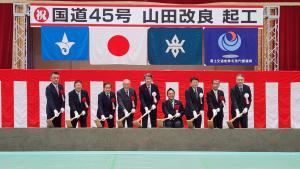 「国道45号山田改良起工」の横断幕の下に紅白幕が張られ、胸章をつけた9名の方が並んで盛り土に鍬入れをしている写真