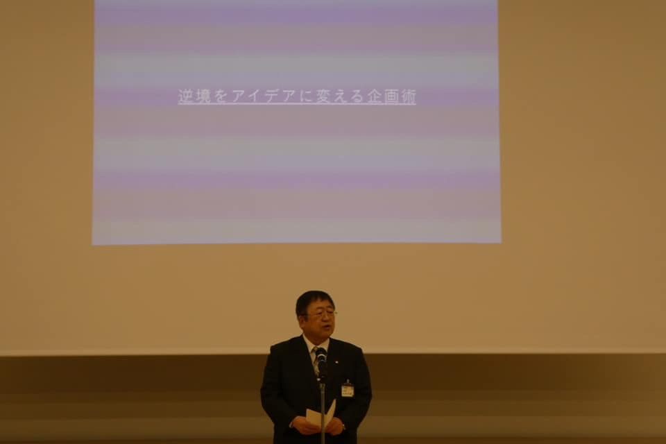 「逆境をアイデアに変える企画術」という文字が投影されたスクリーンの前で、スタンドマイクに向かい話をする様子の市長の写真