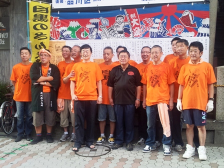 市長とオレンジ色のTシャツを着た目黒駅前商店街振興組合の方々が記念撮影している写真