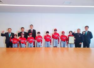 赤いユニフォームを着たサンライズ千徳スポーツ少年団の8人の児童たちと市長と副市長、関係者3人が右手がガッツポーズをしている写真