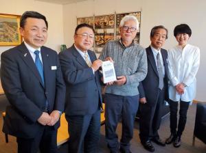 1冊の本を一緒に持つ市長と白髪の男性を中心に、右側に関係者の男性1名、左側に男性と女性が1名ずつ立って並んでいる写真
