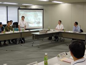 会議室のスクリーンに資料が映し出され、コの字型で並べられた長机の席に関係者が座っており、市長が発言している様子の写真