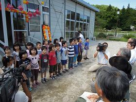 カラフルなペーパーフラワーで飾られた仮設保育所の外で園児たちが横2列に並んでおり、市長や関係者が向かい合わせに座っている写真