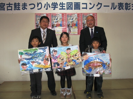 「宮古鮭まつり小学生図画コンクール表彰式」の吊り看板前で、魚が描かれた作品を手に持つ子供たち3人と、市長と澤田克司実行委員会会長が映っている写真