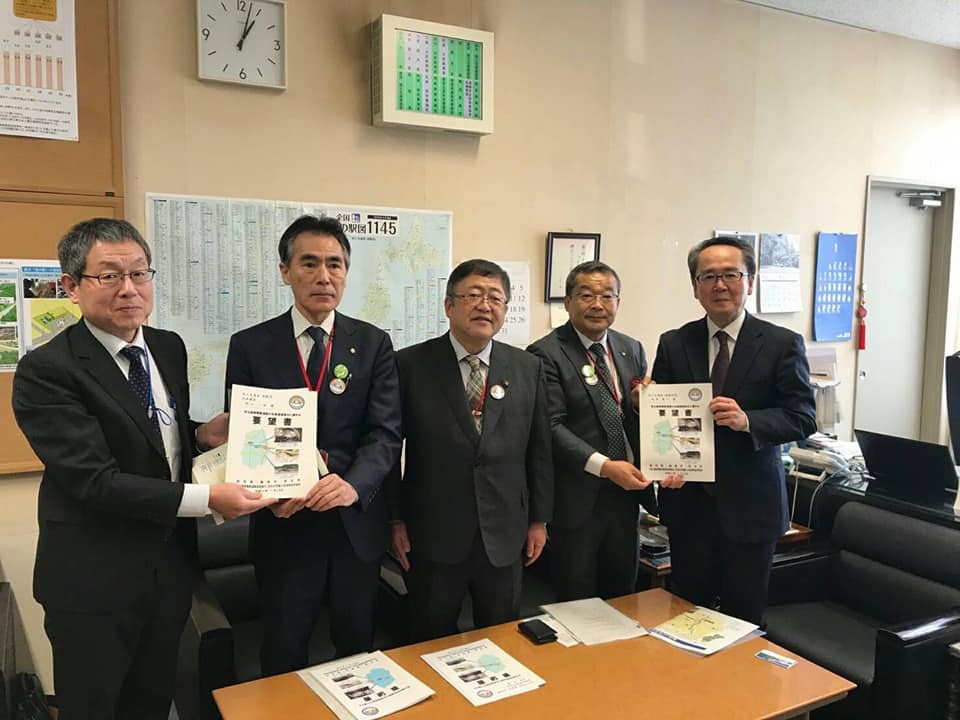 日本地図などの掲示物が貼られた室内で、市長を中央に国土交通省の池田豊人道路局長と村山一弥道路局企画課長、2名の関係者が一緒に要望書を持ち横一列に並んでいる写真