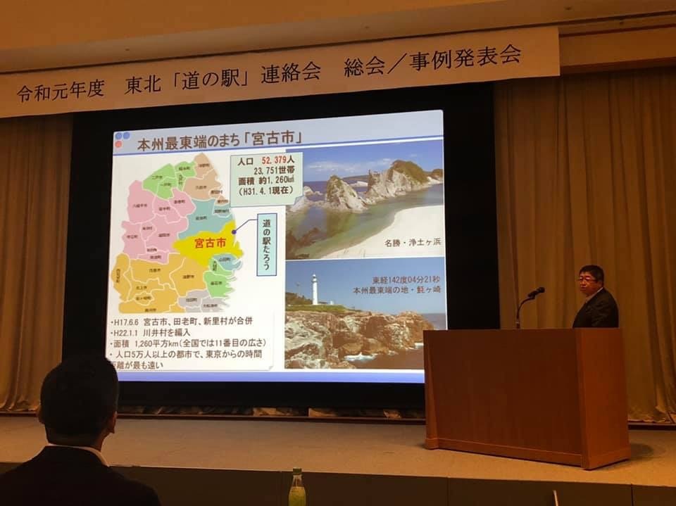 「東北「道の駅」連絡会総会・事例発表会」と書かれた横断幕と資料が映し出されたスクリーンが設置されたステージで、檀上に立つ市長が発表を行っている写真