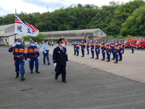横一列に並び直立している消防団員と、制服を着て歩いている市長、市長の後方で旗を掲げて歩く2名の消防団員を写した写真