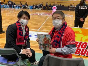 バスケットコートの外側で、ユニフォームを着て首からタオルを掛け両手にチラシのようなものを持った市長と関係者の男性がパイプ椅子に座り、後方を向くように体をひねり一緒に写っている写真