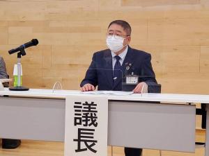 「議長」と大きく書かれた紙が長机の前に貼られパーテーションが設置され資料が置かれた席に座り話を聞いている市長の総会の様子の写真