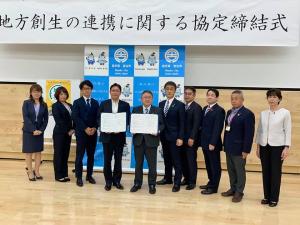 壁の上部に掲げられた「地方創生の連携に関する協定式」と書かれた横断幕と、壁際にバックパネルが置かれた会場で、締結書を正面に向けて両手で広げる男性と市長を中心に、左側に女性2名と男性1名、右側に女性1名と男性4名が並んで写っている写真