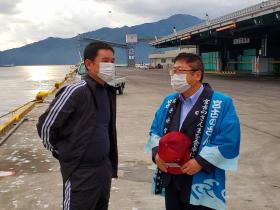 青の法被を着て、赤い帽子を持った市長が黒のジャージ姿の鈴木俊次漁労長と 背景に倉庫が見える宮古港の岸壁付近で話している写真
