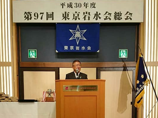 「平成30年度 第97回 東京岩水会総会」と書かれた横断幕と東京岩水会の旗が吊るされた壁の前に置かれた演台のマイクに向かい話をする様子の市長の写真