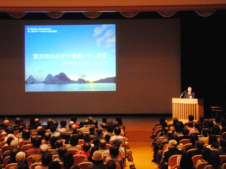 会場のホールに「宮古市は必ずや復興いたします」と題した資料がスクリーンに投影されており、舞台横の演壇で市長が講演する様子を客席側から見た写真