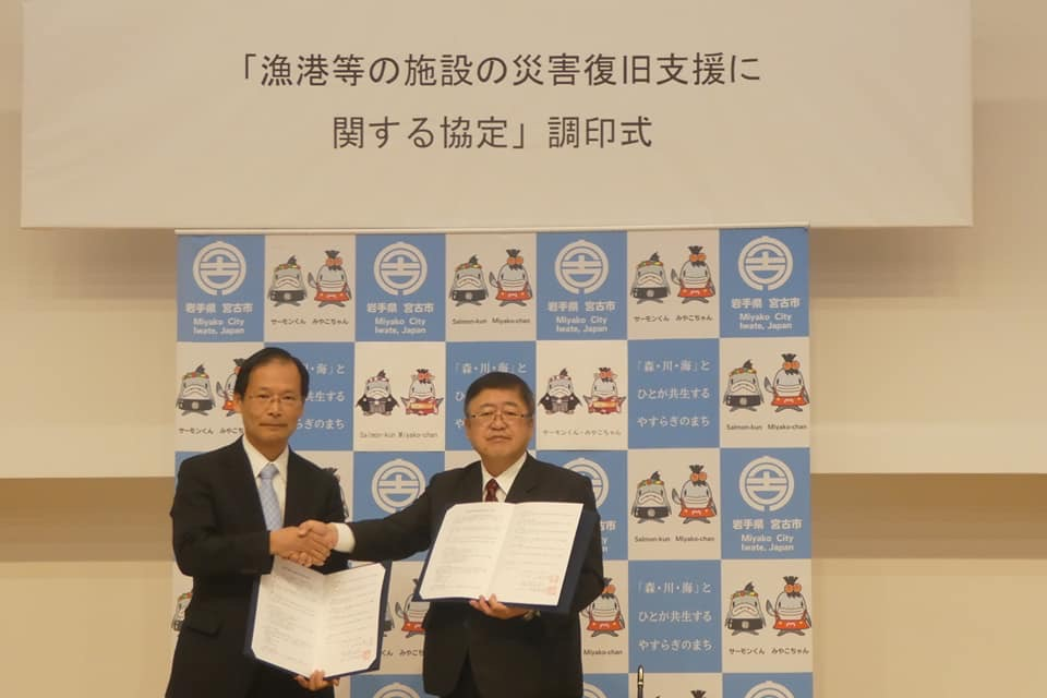 「「漁港等の施設の災害復旧支援に関する協定」調印式」と書かれた横幕と宮古市のバックパネルの前で、一般社団法人水産土木建設技術センターの宇賀神義宣理事長と市長がそれぞれ協定書を開いて持ち、右手で握手を交わしている写真