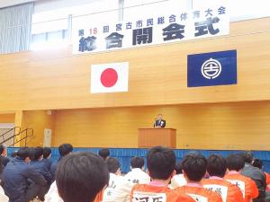 「第18回宮古市民総合体躯大会 総合開会式」と書かれた横断幕が上に掲げられ、その下の演台で話す市長とフロアに座って話を聞く参加者たちの写真