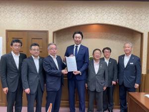 出入り口のドアの前で、中央に財務省の金子俊平政務官と市長会の代表が一緒に要望書を持って中央に立ち、市長を含む5名の関係者が横一列に並んでいる写真