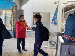 市長が赤の上着に黄色の襷をかけて、改札口を出てきたリュックを背負ったジャージ姿の女性に交通安全運動を行っている写真