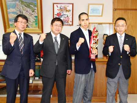応接室で、市長とトロフィーを持った久保田勝幸さんと関係者が4人で並び、右手の拳をあげている写真