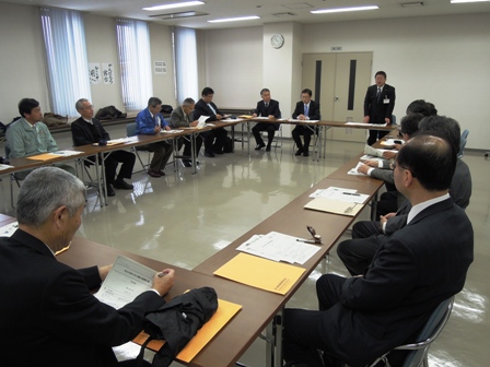 室内で前方にスーツを着た男性が立って話しているのを、口の字型に設置されたテーブルと椅子に座り話を聞いている関係者の写真