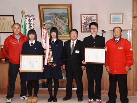 市長を中心に、トロフィーを持つ女子学生、賞状を持つ女子学生と男子学生、赤いジャケットを着た2名の男性が横一列に並ぶ記念写真