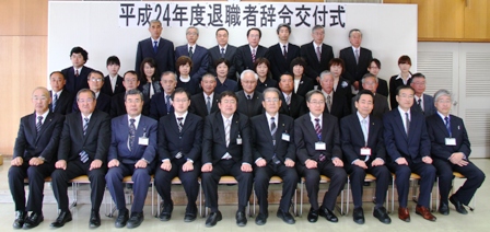 「平成24年退職者辞令交付式」と書かれたバックパネルの前で、市長と関係者らが横4列に並んでいる写真