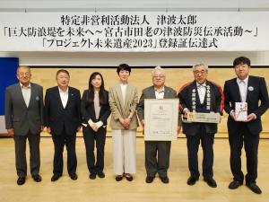 式の題名が書かれた横断幕が吊るされた会場で、認定証や目録を持つ関係者や市長が横一列に並ぶ記念写真
