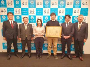 バックパネルの前で賞状の額縁を持つ男性と記念品を持つ女性を中心に、市長と関係者の男性1名が左側、関係者の男性2名が右側に並んで立っている記念写真