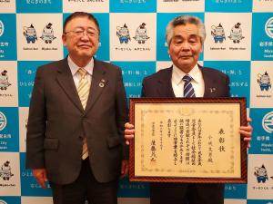 バックパネルの前で賞状の額縁を持つ小成文男さんと市長が並んで写っている写真