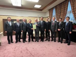 国旗や白い応接セットが設置された室内で、財務大臣の鈴木俊一氏と市長が一緒に要望書を持って中央に立ち、8名の県内首長の方々が横一列に並んでいる写真