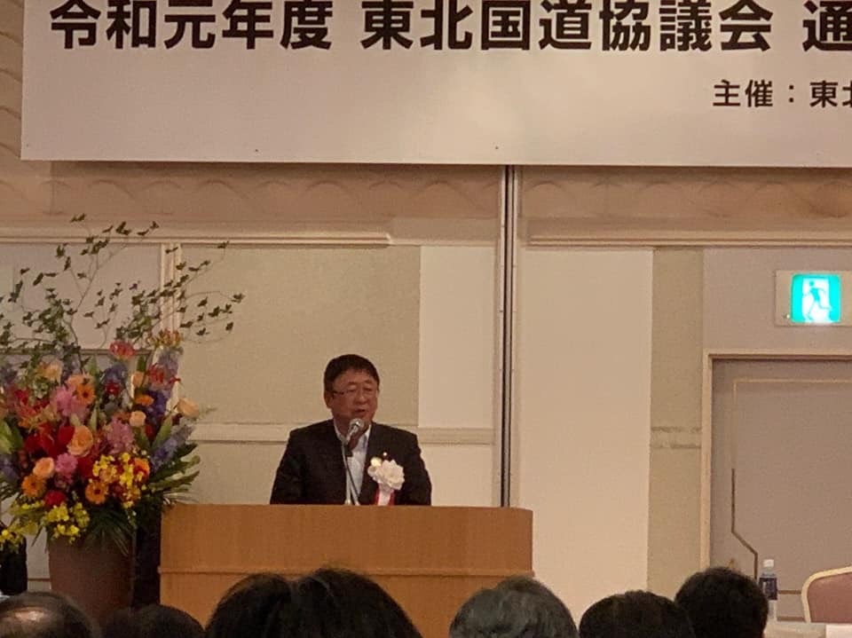 カラフルな色の花が生けられた演台花の傍に設置された檀上で、白色の胸章リボンを付けた市長が挨拶を行っている写真