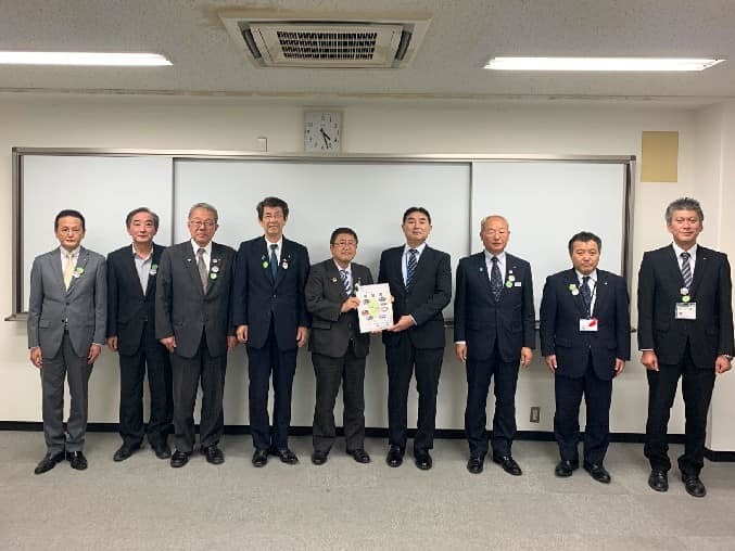 ホワイトボードの前で、要望書を一緒に持つ市長と男性、7名の関係者の男性が横一列に並ぶ記念写真