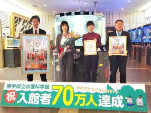 「入館者70万人達成」と書かれた看板の後方で額に入ったポスターを持った男性、花束を持った女性と額に入った賞状を持った女性、額縁を持った市長が横一列に並んでいる記念写真