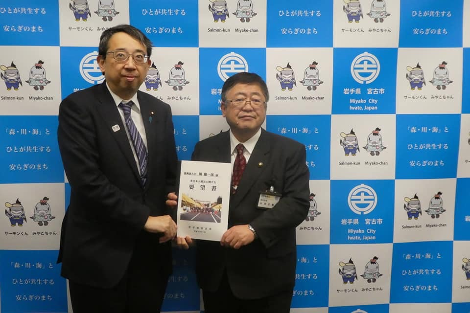 宮古市のバックパネルの前で、橘慶一郎復興副大臣と市長が一緒に要望書を持ち立っている写真