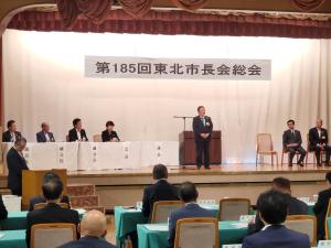 「第185回東北市長会総会」と書かれた横断幕が吊るされたステージ上の演台の両脇に関係者が座り、ステージ下には参加者の方々が座っている中、演台の前で市長が話をしている写真