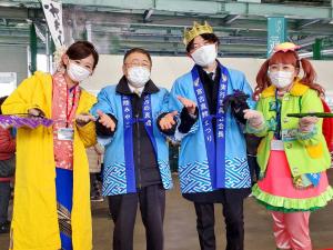 青い法被を着た市長、青い法被を着て頭に冠を載せた男性を中心に、黄色い衣装をきた女性が左側、右側にピンクと黄緑の衣装を着て髪を耳の上で二つ結びにしている女性が写っている写真