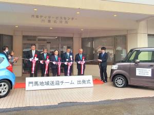 「門馬地域チーム出発式」と書かれたボードが置かれた後方で、送迎サービスで使用する車の間で市長を含む5名でテープカットをしている写真