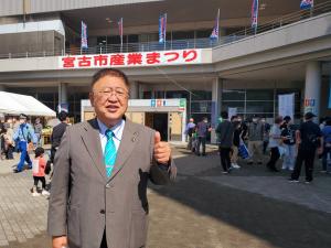 体育館の壁に「宮古市産業まつり」と書かれた横断幕が掛けられ、その体育館の前で左手の親指を上にむけポーズをとる市長を写した写真