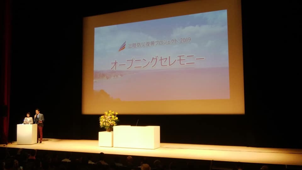「三陸防災復興プロジェクト2019オープニングセレモニー」という文字と海の写真が投影されたスクリーンが設置された舞台で、演台の前に立つ女性1名と男性1名の写真