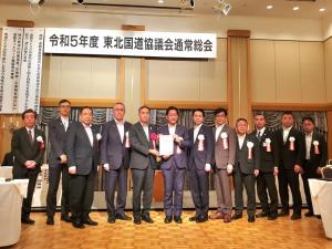 「令和5年度 東北国道協議会通常総会」と書かれた看板が掲げられ、その下でスーツを着て胸章をつけた男性12人が横並びで写っている写真