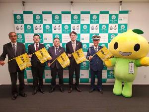 宮古市のバックパネルの前で、制服姿の男性と市長、3名の関係者が寄贈された黄色の横断旗を持ち、こくみん共済coopのキャラクター「ピットくん」と横一列に並んでいる写真