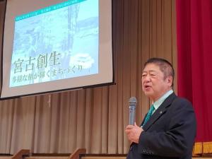 「宮古創生」と書かれた画像が映し出されたスクリーンの横で、市長がマイクを持って話をしている写真
