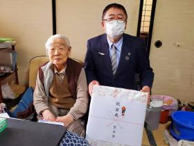 上林ハルさんのご自宅の居間で市からの記念品を持った市長と上林ハルさんが座って撮影した写真
