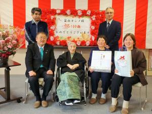 前列には市長、車いすに乗った女性、記念品を持つ2人の女性が椅子に座り、後列に立つ2人の男性を写した写真