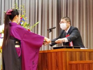 紫色の袴を着た女性が、演台の向かい側から市長に賞状を両手で手渡されている写真