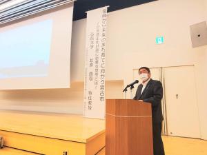 前方に資料が映し出されたスクリーンが設置され、右側の檀上で挨拶を行っている市長の写真