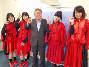 市長を中心に、真っ赤なスカートの衣装を身に着けた女性が両端に2名ずつ並び、全員が右手の親指と人差し指を合わせて丸の形を作りポーズを決め笑顔で写っている写真