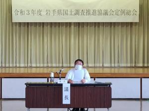 令和3年度岩手県国土調査推進協議会定例総会の横断幕の下で、議長と書かれた紙が貼られた机で市長が資料を見ている写真