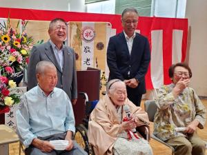 紅白幕の前に花が飾られた場所で、車椅子に乗っている久保田イチさんがマイクで話をされて、両脇に座っている男女、後ろに立っている市長と男性が笑っている写真
