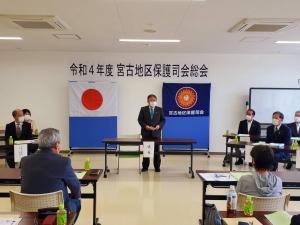 「令和4年度宮古地区保護司会総会」と書かれた横断幕と、国旗と会旗が貼られた壁の前で立って話す様子の市長と、市長を囲むように置かれたテーブルに着席している関係者の方たちの写真