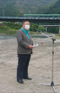 川岸に置かれたスタンドマイクの前で両手で紙を持ち話をしている様子の市長の写真